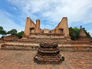 Fototapeta premium Ancient Ruins in Thailand: Sun-drenched ancient ruins in Thailand, featuring weathered brick structures and pagodas, stand testament to a bygone era under a clear blue sky dotted with fluffy clouds.