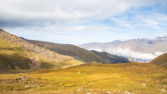 A golden alpine plateau scattered with rocks overlooks layered ridges and clouds toward distant Mount Kazbek, Georgia.