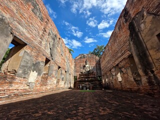 Fototapeta premium Ancient Buddhist Temple Ruins: The weathered brick walls of an ancient temple stand against a vibrant blue sky, with a serene Buddha statue at its center.
