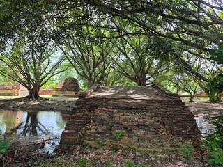 Fototapeta premium Ancient Ruins in Lush Vegetation: Weathered brick ruins, likely part of an old structure, emerge from overgrown vegetation beside a tranquil body of water, framed by the shade of majestic trees.