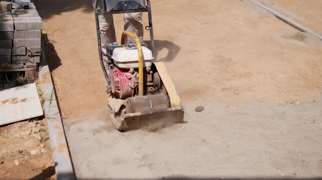 Worker operates plate compactor on unfinished dirt beside stacked paving blocks, while dust rises from surface preparation and rough ground compacts under bright sunlight