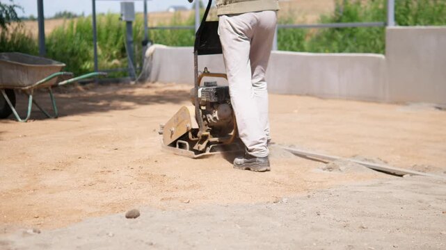 Low-angle construction scene shows worker legs guiding plate compactor across dusty prepared ground at outdoor building site, with visible vibration, disturbed soil, and nearby