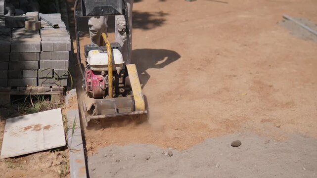Compacting machine stands beside stacked gray paving blocks on dusty earth, with worker legs behind it, wheelbarrow in background, and open sunlit ground at right creating