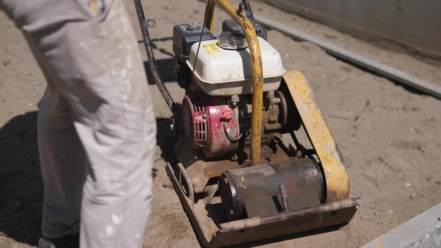 Construction worker at left foreground guiding compact plate compactor across dusty outdoor worksite, with worn yellow frame, white tank, red engine housing, rust, hard shadows