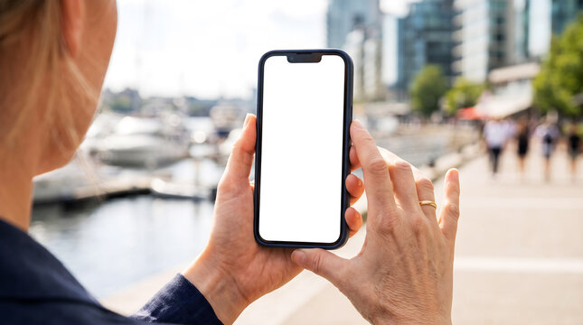 Hand holding mobile phone with blank white screen mockup at city waterfront marina, blonde woman on promenade using yacht charter and harbor navigation app, travel concept
