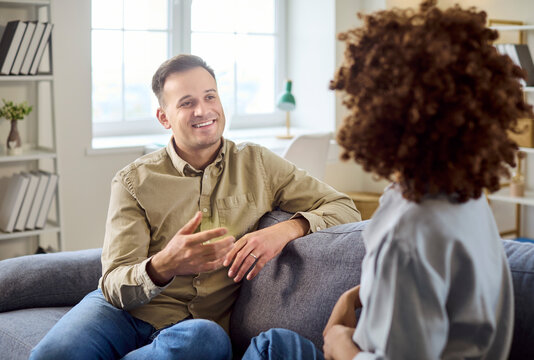 talking on sofa at home, conversation. Smiling man listens to woman in a bright living room, showing relationship trust and empathy. Clear concept is communication at home.