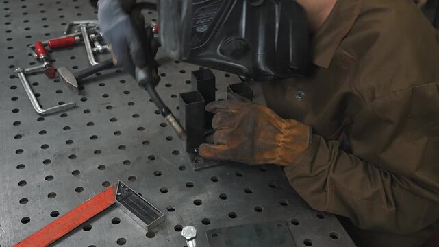 A working engineer TIG welds metal structural parts. He wears a protective mask and gloves to avoid injury from hot sparks