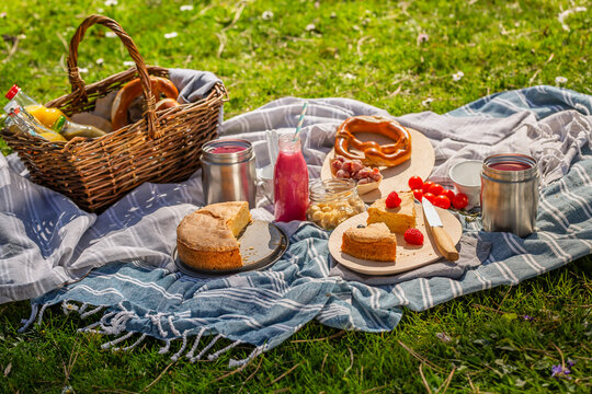 A woven basket filled with beverages and treats sits on a checkered blanket laid out on lush green grass