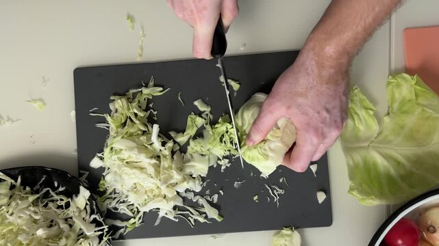 Close view of hands slicing pale green cabbage on dark cutting board with knife pressing through firm wedge as thin shreds gather nearby, showing food prep in domestic kitchen with natural light and