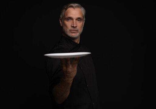 Portrait Of 50s Mature Chef In Uniform Holding Empty Plate on black.