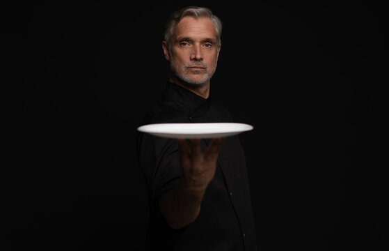 Portrait Of 50s Mature Chef In Uniform Holding Empty Plate on black.