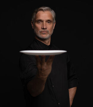 Portrait Of 50s Mature Chef In Uniform Holding Empty Plate on black.