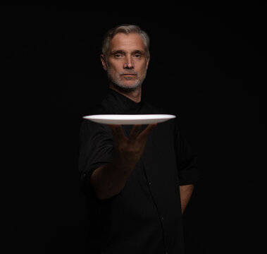 Portrait Of 50s Mature Chef In Uniform Holding Empty Plate on black.