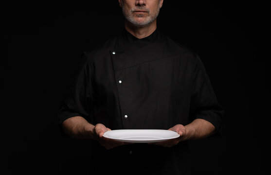 Portrait Of 50s Mature Chef In Uniform Holding Empty Plate on black.