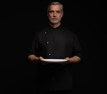 Portrait Of 50s Mature Chef In Uniform Holding Empty Plate on black.