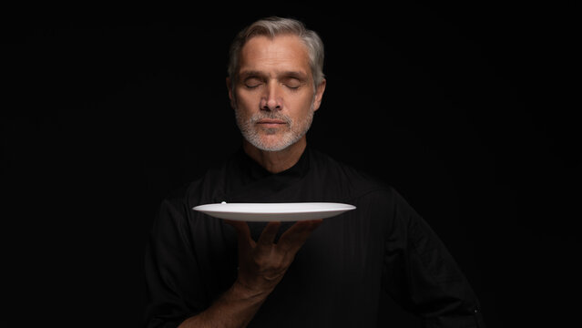 Portrait Of 50s Mature Chef In Uniform Holding Empty Plate on black.