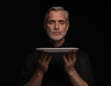 Portrait Of 50s Mature Chef In Uniform Holding Empty Plate on black.