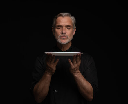 Portrait Of 50s Mature Chef In Uniform Holding Empty Plate on black.