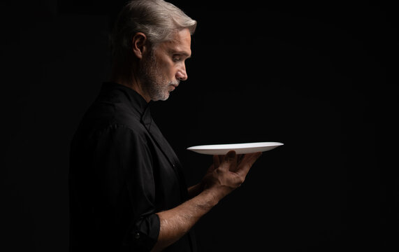 Portrait Of 50s Mature Chef In Uniform Holding Empty Plate on black.