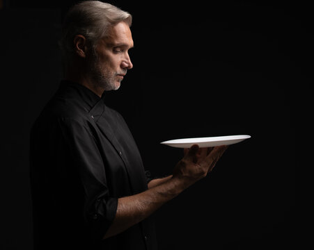 Portrait Of 50s Mature Chef In Uniform Holding Empty Plate on black.