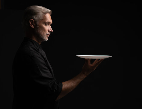 Portrait Of 50s Mature Chef In Uniform Holding Empty Plate on black.