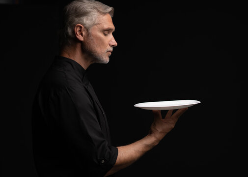 Portrait Of 50s Mature Chef In Uniform Holding Empty Plate on black.