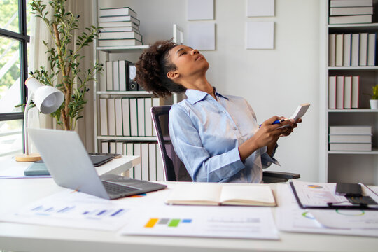 Young businesswoman relaxing in an office chair, holding a calculator while taking a break from working on her laptop and reviewing business charts and documents.