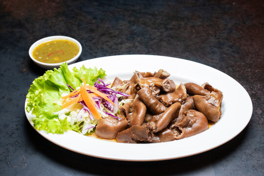 Stewed pork chitterlings in brown sauce served with spicy dipping sauce and vegetables on white plate over dark background.