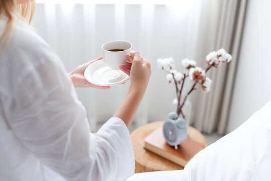 Woman enjoying morning coffee in bed. Slow living, self-care, relaxation and mindfulness routine concept. Home interior with alarm clock, lifestyle moment, close up