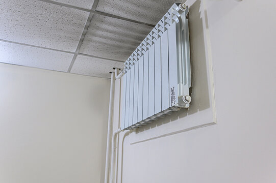white 15‑section bimetal radiator is mounted on plain wall. connected via vertical white pipes, beneath speckled acoustic ceiling tile grid
