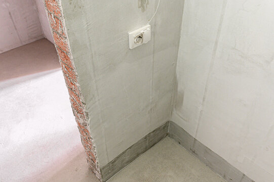 corner with rough‑plastered walls features exposed brickwork along edge. white Legrand‑style electrical outlet is mounted mid‑wall, with grey skirting board and plain concrete floor