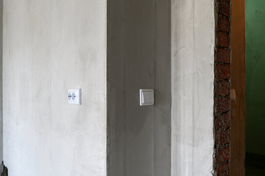 light‑plastered wall hosts white double socket and single light switch. grey door jamb aligns with textured plaster edge, while exposed red‑brown brickwork frames adjacent doorway