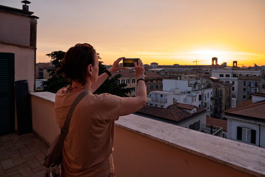Woman standing on a rooftop terrace taking a photo of a beautiful golden Rome sunset over the eternal city skyline with her smartphone