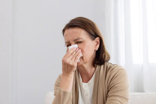 Mature woman sneezing with tissue sitting on a couch feeling unwell in living room