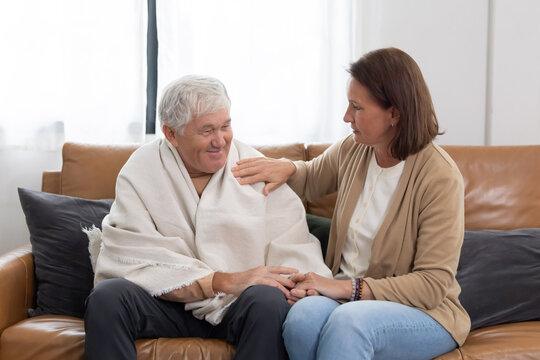 Elderly man sits on couch with woman who comforts him while he is wrapped in a blanket