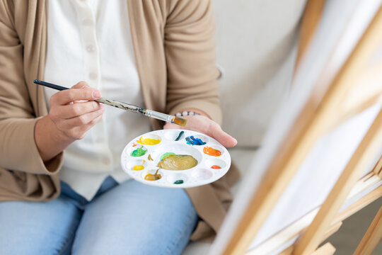 Person holds a paintbrush above a palette with colors while sitting in front of an easel