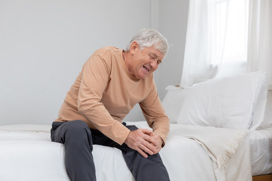 Older man sitting on bed holding his knee in pain during morning