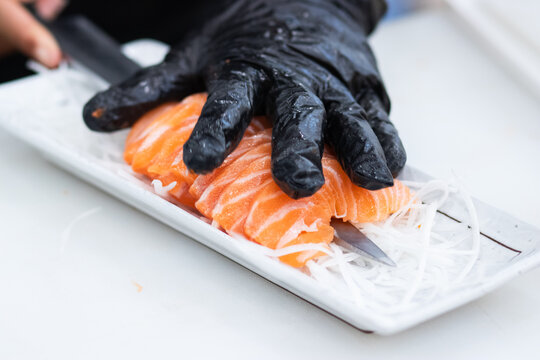 A sushi chef in black gloves transfers fresh raw salmon sashimi slices with a kitchen knife onto a white ceramic plate with daikon radish for gourmet Japanese dining nutrition.