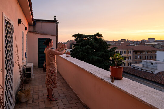 Side view of a woman standing on a long rooftop balcony in Rome, taking a smartphone photo of the golden sunset over the eternal city skyline