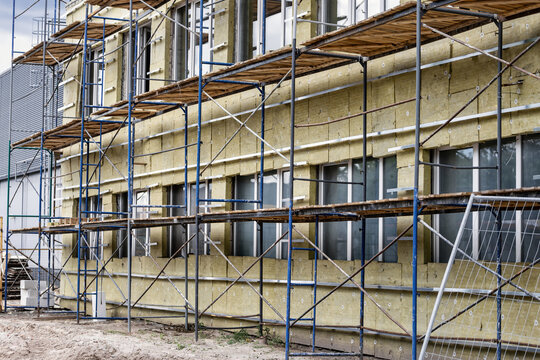 Workers use scaffolding to insulate the facade of a building with mineral wool. The site shows construction activity and safety measures in place