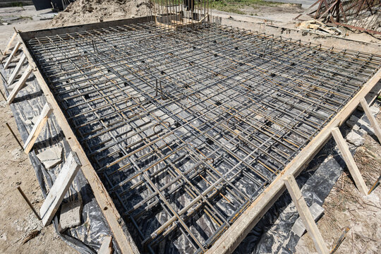 Workers set up rebar mesh in a foundation area for concrete pouring. The site has a flat surface and wooden forms surrounding the rebar