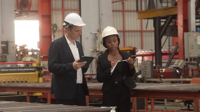 Medium shot of a Caucasian male executive and a Black female manager wearing safety helmets while reviewing operational data on a digital tablet in a heavy industrial plant. Concept of corporate 