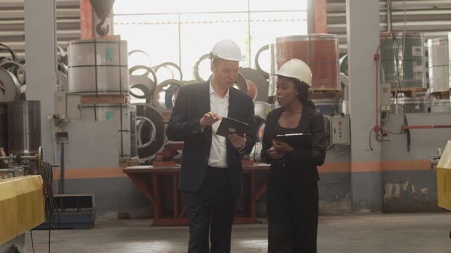 Medium shot of a Caucasian male executive and a Black female manager wearing safety helmets while reviewing operational data on a digital tablet in a heavy industrial plant. Concept of corporate 