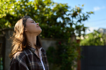 Fototapeta premium Smiling woman with shoulder length hair standing outdoors, head tilted toward the sun. Casual plaid shirt, nature backdrop, feeling relaxed, carefree, and reflective