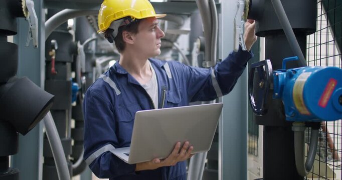 An industrial engineer in a hard hat uses a laptop for diagnostics while inspecting complex machinery in a factory, ensuring system optimization and quality control.