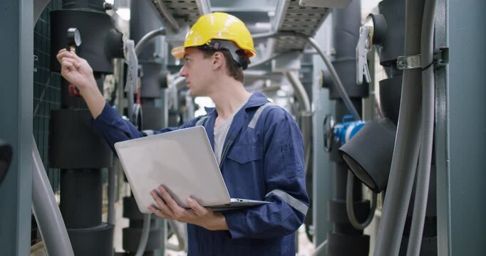 An industrial engineer in a hard hat uses a laptop for diagnostics while inspecting complex machinery in a factory, ensuring system optimization and quality control.