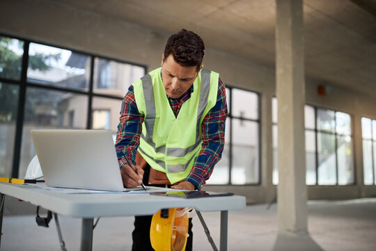 Construction worker using a laptop and examining architectural blueprints inside a building under construction.