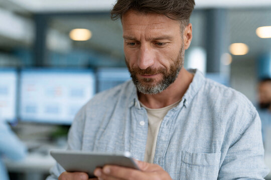 Focused man studies data on a tablet in an office. His brow furrows slightly in concentration. Background screens display business metrics. He wears a casual, light-colored button-up shirt