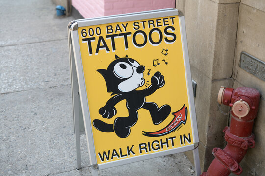 a-frame sidewalk sign outside 600 Bay St featuring a cartoon cat whistling towards a tattoo shop, Toronto