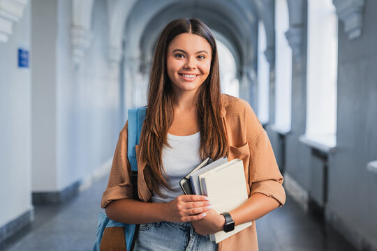 Female student with backpack holding books and notebooks in school corridor looking at camera, confident teenager, study, education and school life concept